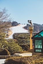 A snow-covered ski slope with numerous people skiing down, surrounded by evergreen trees. In the foreground, a whimsical structure resembling a pirate-themed lookout tower with a skull and crossbones flag is visible. A modern glass gazebo is positioned to the right. The background features a clear blue sky and distant tree-covered hills.