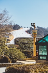 A snow-covered ski slope with numerous people skiing down, surrounded by evergreen trees. In the foreground, a whimsical structure resembling a pirate-themed lookout tower with a skull and crossbones flag is visible. A modern glass gazebo is positioned to the right. The background features a clear blue sky and distant tree-covered hills.