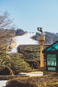 A snow-covered ski slope with numerous people skiing down, surrounded by evergreen trees. In the foreground, a whimsical structure resembling a pirate-themed lookout tower with a skull and crossbones flag is visible. A modern glass gazebo is positioned to the right. The background features a clear blue sky and distant tree-covered hills.