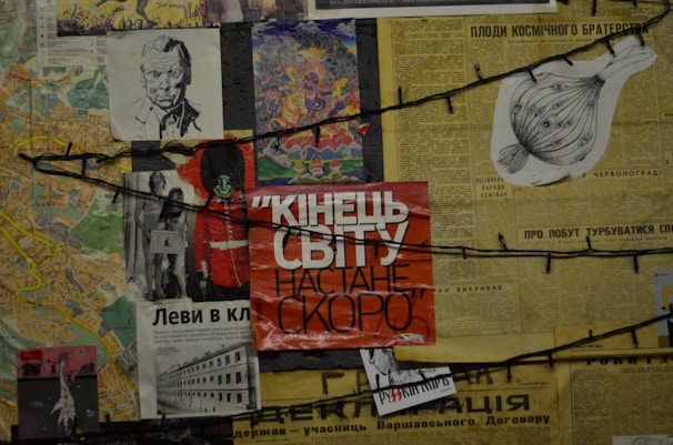A collage of various newspaper clippings and images, including a large red poster with Cyrillic text, a black and white sketch of a man, and colorful illustrations. Strings run across the collage, adding a chaotic and layered effect.