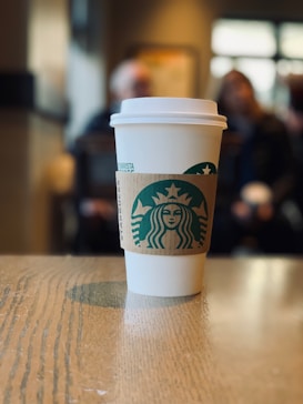 A branded coffee cup with a cardboard sleeve sits on a wooden table. The background is blurred, showing out-of-focus people and a window, creating a cozy indoor atmosphere.
