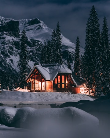 A cozy cabin is illuminated warmly amidst a snowy landscape. Tall pine trees surround the cabin, and a majestic, snow-covered mountain provides a stunning backdrop under a dark, cloudy sky.