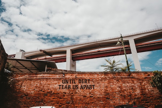 A brick wall with the phrase 'UNTIL DEBT TEAR US APART' painted on it. Above the wall, there is a large concrete bridge with red accents. A green lamp post and some foliage are visible in front of the wall. The sky is partly cloudy.