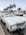 A white classic convertible car parked on the side of a street lined with tall palm trees. Other cars are driving on the road, and there are various storefronts visible in the background. The sky is clear, giving a bright and sunny atmosphere.