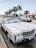 A white classic convertible car parked on the side of a street lined with tall palm trees. Other cars are driving on the road, and there are various storefronts visible in the background. The sky is clear, giving a bright and sunny atmosphere.