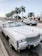 A white classic convertible car parked on the side of a street lined with tall palm trees. Other cars are driving on the road, and there are various storefronts visible in the background. The sky is clear, giving a bright and sunny atmosphere.