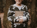 A child wearing a camouflage sweater is outdoors, holding a book titled 'Our Bird Friends' with arms crossed in front. The background features tree trunks and a natural forest environment.