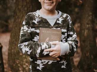 A child wearing a camouflage sweater is outdoors, holding a book titled 'Our Bird Friends' with arms crossed in front. The background features tree trunks and a natural forest environment.
