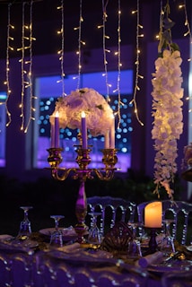 A beautifully decorated table is set with a golden candelabra holding several white candles and a floral arrangement. Hanging string lights provide a warm, enchanting ambiance. To the right of the candelabra, there is a cascading white floral arrangement. A lit pillar candle is placed on the table near intricately folded napkins and upside-down wine glasses. The background is illuminated with purple and blue hues, adding a dreamy effect.