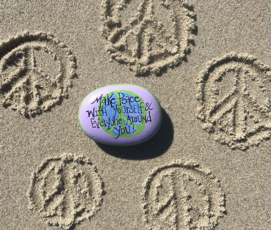 A painted rock rests on sandy ground, surrounded by multiple peace symbols etched into the sand. The rock is colored with a purple and green design, resembling the Earth, and bears the handwritten message 'Make Peace With Yourself & Everyone Around You'.