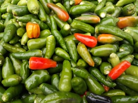 A pile of assorted chili peppers, predominantly green with a few orange and red ones interspersed throughout. The peppers are shiny and appear fresh, with varying shapes and sizes.