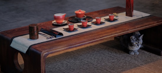 A wooden table is set with a traditional tea set, including a teapot, cups, and a serving tray, all in a red hue. The setup exudes a simplistic and elegant aesthetic, laid out on a table runner. A gray cat sits quietly underneath the table on a woven mat.