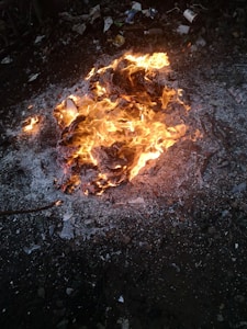 A fire burning among debris and ash on the ground with visible orange flames. Surrounding the fire, there are pieces of charred material, litter, and scattered garbage.