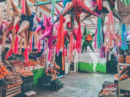 Colorful piñatas hang from the ceiling in a lively market setting, surrounded by an array of fresh fruits like oranges, bananas, and pineapples. The scene includes a child's riding toy shaped like a horse.
