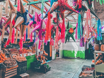 Colorful piñatas hang from the ceiling in a lively market setting, surrounded by an array of fresh fruits like oranges, bananas, and pineapples. The scene includes a child's riding toy shaped like a horse.