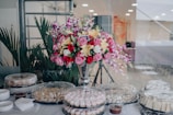 A large bouquet of assorted flowers, including roses and lilies, is arranged in a tall, elegant vase. The bouquet is surrounded by a spread of covered dishes, likely containing various pastries and desserts, and is set on a white tablecloth. The setting appears to be indoors with green potted plants in the background.