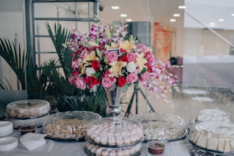 A large bouquet of assorted flowers, including roses and lilies, is arranged in a tall, elegant vase. The bouquet is surrounded by a spread of covered dishes, likely containing various pastries and desserts, and is set on a white tablecloth. The setting appears to be indoors with green potted plants in the background.