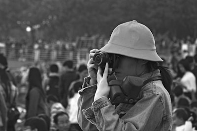 A person wearing a hat and denim jacket is using a camera to take a photograph amidst a large crowd. There is a pair of headphones around their neck, and the setting appears to be outdoors with blurred people in the background.
