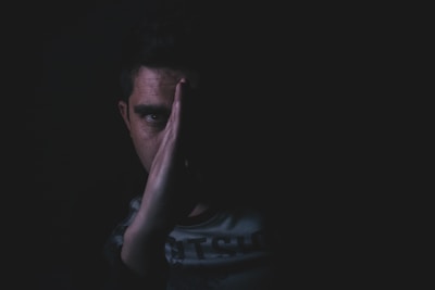 A person is partially obscured by shadows, with one side of their face visible and the other hidden. Their hand is pressed against their face, creating a mysterious and introspective appearance. The lighting is low, emphasizing the contrast between light and shadow.