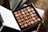 A box of assorted chocolates neatly arranged in rows, with varying shapes, textures, and embellishments like gold leaf. The box is open and placed on a surface with a soft focus background.
