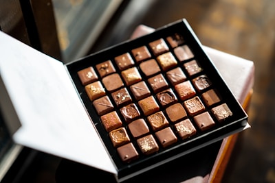 A box of assorted chocolates neatly arranged in rows, with varying shapes, textures, and embellishments like gold leaf. The box is open and placed on a surface with a soft focus background.