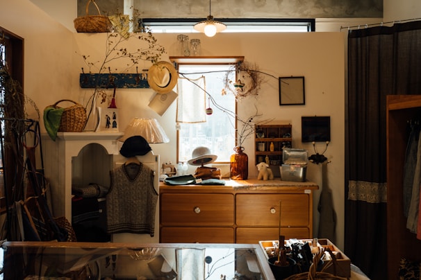 A cozy, well-decorated interior space with a mix of vintage and rustic elements. The room includes a wooden dresser topped with assorted items like a glass vase, a small ceramic bear, and a wicker basket. A cozy knitted vest is displayed on a white cabinet, and a collection of hats are hung nearby. Various decorative plants and branches provide a natural touch, while a lamp with a unique angled shade adds to the lighting. A window at the top allows natural light to filter in, contributing to the warm ambiance.