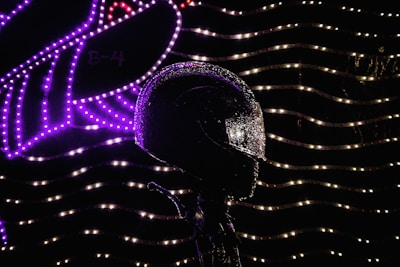 A motorcycle helmet is covered in water droplets and illuminated by bright, colorful lights in the background, arranged in wavy patterns with purple and white hues.