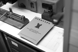 A black and white photograph featuring an industrial printing machine setup. A booklet titled 'LINOTYPE' is prominently displayed on the table, alongside other machinery components. The scene evokes a sense of a traditional printing press environment.
