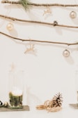 A minimalist holiday decoration setup featuring pine branches, small pinecones, and gold ornaments hanging from thin wooden sticks against a white background. Below, there is a candle inside a glass holder with moss, and a pinecone resting on the surface.