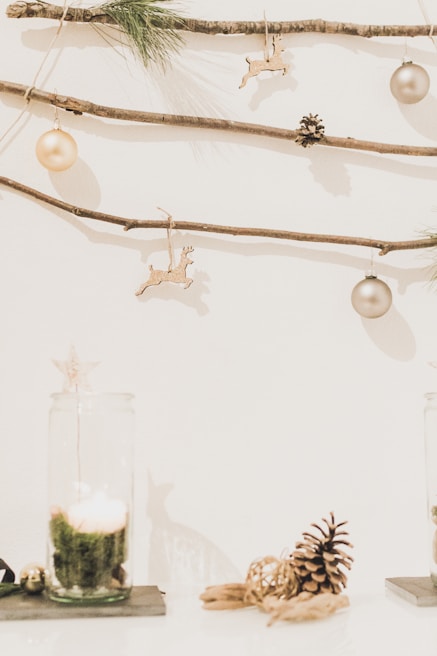 A minimalist holiday decoration setup featuring pine branches, small pinecones, and gold ornaments hanging from thin wooden sticks against a white background. Below, there is a candle inside a glass holder with moss, and a pinecone resting on the surface.