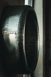 A close-up view of a large metallic, cylindrical structure with rivets and a shiny, reflective surface. The object appears industrial, possibly part of machinery or an aircraft component. The lighting creates a dramatic contrast, highlighting the smooth, curved contours of the metal.