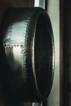 A close-up view of a large metallic, cylindrical structure with rivets and a shiny, reflective surface. The object appears industrial, possibly part of machinery or an aircraft component. The lighting creates a dramatic contrast, highlighting the smooth, curved contours of the metal.