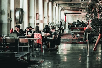 A spacious, industrial-style interior room features multiple people sitting at tables, engaging in activities like using laptops and conversing. The floor is polished concrete, and plants are strategically placed, adding greenery to the otherwise minimalistic decor. Large windows line one wall, allowing natural light to enter the space, and there are decorative elements like area rugs and wall art present.