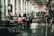 A spacious, industrial-style interior room features multiple people sitting at tables, engaging in activities like using laptops and conversing. The floor is polished concrete, and plants are strategically placed, adding greenery to the otherwise minimalistic decor. Large windows line one wall, allowing natural light to enter the space, and there are decorative elements like area rugs and wall art present.