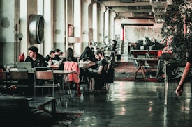 A spacious, industrial-style interior room features multiple people sitting at tables, engaging in activities like using laptops and conversing. The floor is polished concrete, and plants are strategically placed, adding greenery to the otherwise minimalistic decor. Large windows line one wall, allowing natural light to enter the space, and there are decorative elements like area rugs and wall art present.