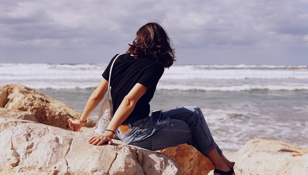 woman in black shirt and blue jeans sitting on rocks by the beach