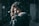 A person is pointing a gun directly at the camera in a dark, possibly snowy or misty, environment. The focus is on the barrel of the gun, which is centered in the image, while the background including the person's face is out of focus.