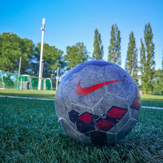 A close-up view of a worn-out soccer ball with a red Nike logo, positioned on a grassy soccer field. In the background, there are goal posts and tall trees under a clear blue sky.