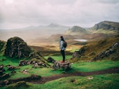 A person stands on a rock overlooking a vast landscape featuring rolling green hills and rocky outcrops with a distant loch and misty mountains under a cloudy sky.