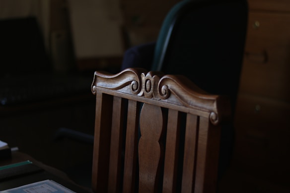 A wooden chair with intricate carvings on the top rail is positioned in a dimly lit room. Behind the chair, an office chair with a dark backrest can be seen, along with a wooden cabinet and a desk.
