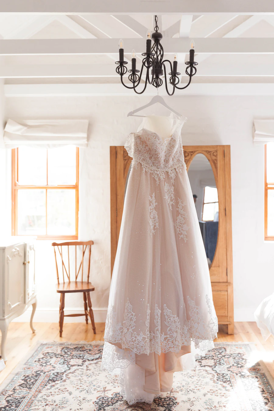 A lace wedding gown hanging from a chandelier in a bright, white room