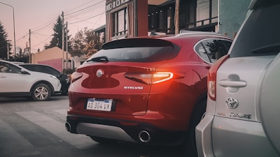 A red Alfa Romeo Stelvio is parked on the street, surrounded by other cars. The rear of the vehicle is prominently visible, with modern designed tail lights and a polished exterior. It's positioned between a white vehicle on the left and a silver Toyota RAV4 on the right. Urban elements like buildings and power lines are in the background.