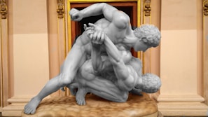 Two intricately carved stone statues of muscular men engaged in a wrestling pose. The figures are positioned on a textured pedestal, set against a richly decorated architectural background featuring ornate gold and tan detailing.