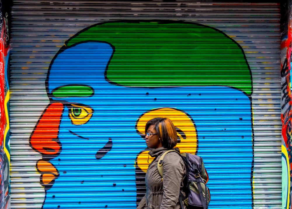Mujer con chaqueta gris caminando cerca de pintura mural azul, naranja y verde