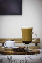 A tall glass mug filled with latte topped with a layer of foam is placed on a wooden board. Next to it is a smaller white cup with a saucer, containing espresso. Coffee beans are scattered around the cups on the board. A wooden bench is visible in the background, creating a cozy cafe ambiance.