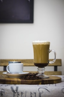 A tall glass mug filled with latte topped with a layer of foam is placed on a wooden board. Next to it is a smaller white cup with a saucer, containing espresso. Coffee beans are scattered around the cups on the board. A wooden bench is visible in the background, creating a cozy cafe ambiance.