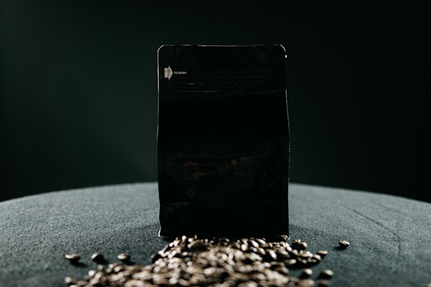 A black package stands upright on a dark surface, surrounded by a scattering of coffee beans in the foreground, with soft, diffused lighting creating dramatic shadows and a minimalist aesthetic.