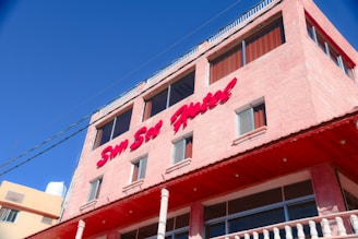 A multi-story building with a light pink exterior and large windows. The building features a prominent sign in red text reading 'Sun Set Hotel'. It has a flat roof with a railing and the sky is a clear shade of blue.
