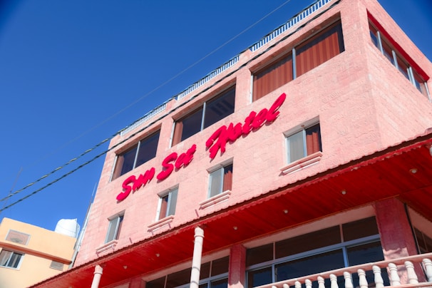 A multi-story building with a light pink exterior and large windows. The building features a prominent sign in red text reading 'Sun Set Hotel'. It has a flat roof with a railing and the sky is a clear shade of blue.