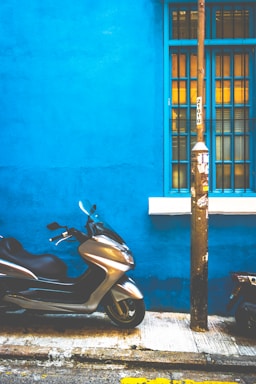 A silver scooter is parked on a sidewalk next to a vibrant blue wall. The wall features a window with blue frames and bars, and there is a utility pole nearby with some notices attached.